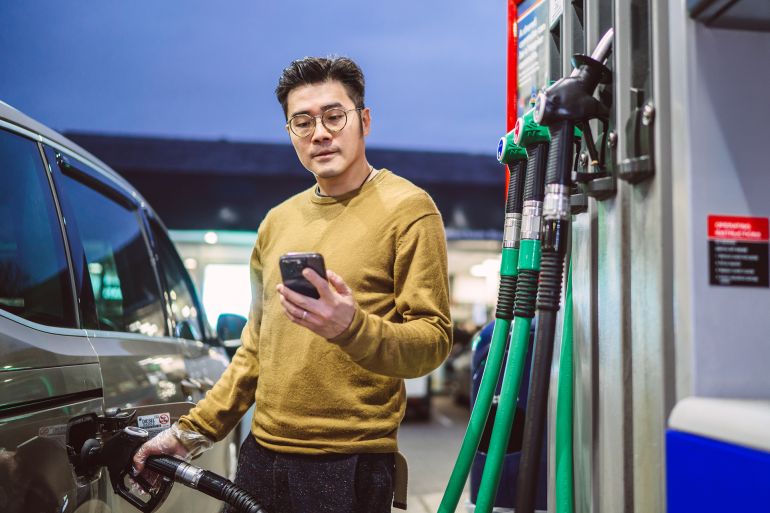 Confident man refuelling his car at a petrol station while checking fuel price on his smart phone. Concept of transportation, energy prices, travel planning, and fuel economy.